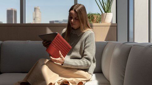 37A27FD6A_Panel5_3upBoxShadow_YourWay.jpg A woman sits on a couch opening Surface Pro 7 with a Poppy Red Type Cover