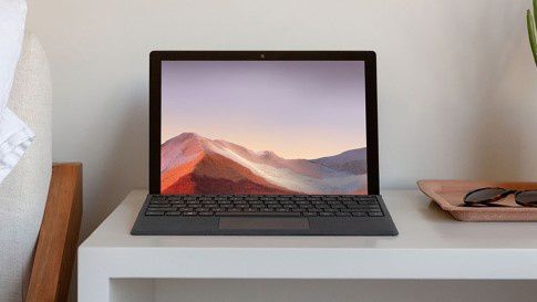 37A27FD6A_Panel8_3upBoxShadow_Display.jpg Surface Pro 7 with a Black Signature Type Cover open on a desk next to a bed