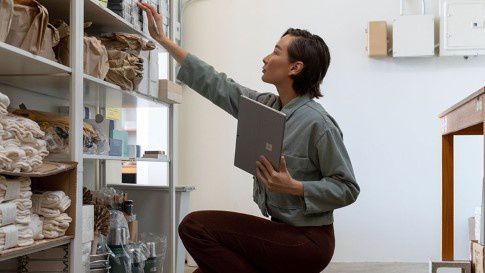 A woman holds Surface Pro 7 while reaching up to a bookshelf