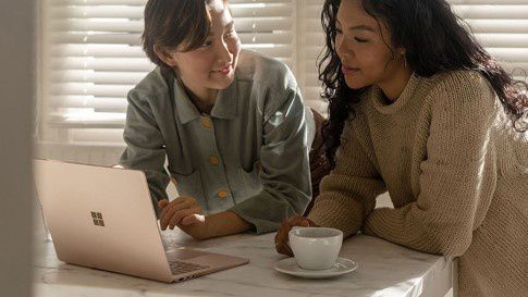 Two women sit at a dining table in front of a Surface Laptop 3