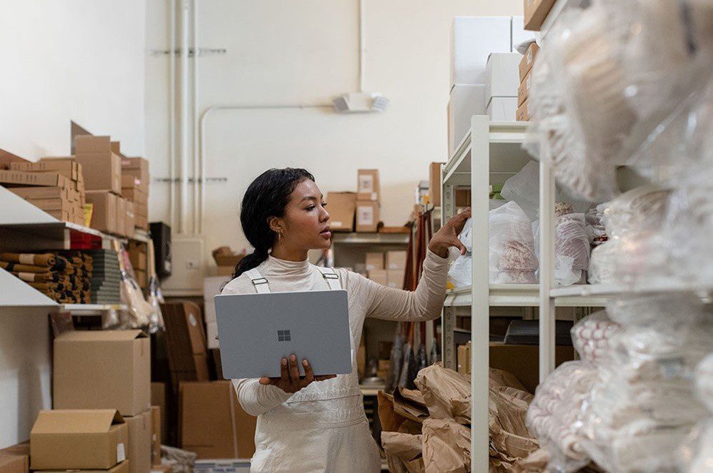 A woman holds Surface Laptop 3 in her hand while reaching for an item on a shelf