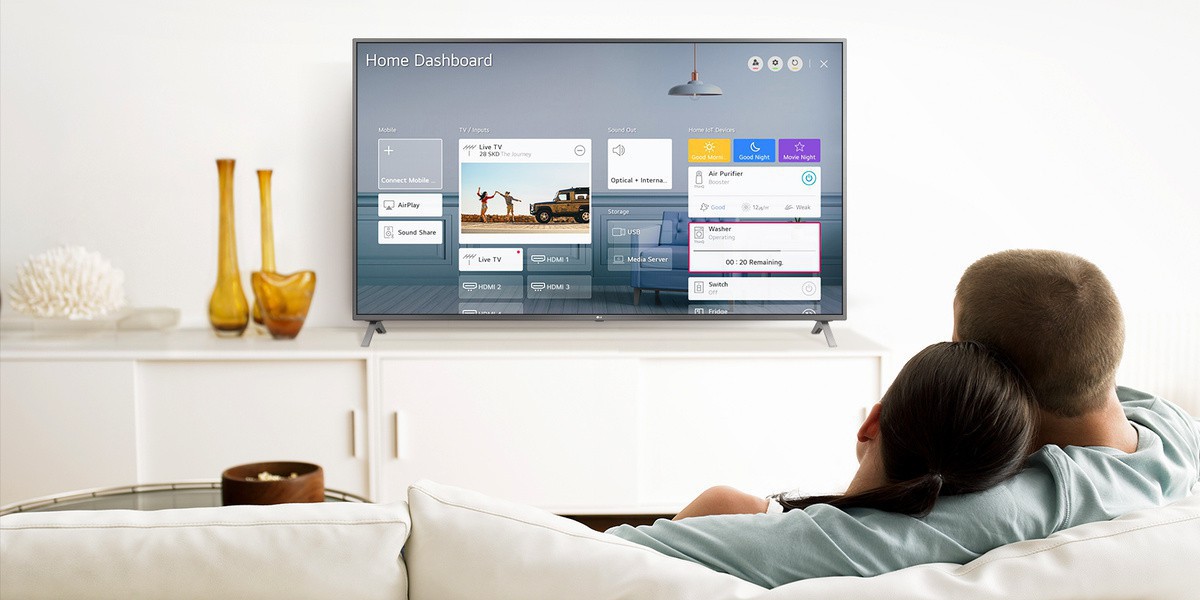 Un bărbat si o femeie care stau pe o canapea în camera de zi, cu Home Dashboard pe ecranul TV.